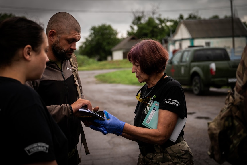 A woman with red hair, a clipboard under her arm, wearing blue surgical gloves hands paperwork to two people