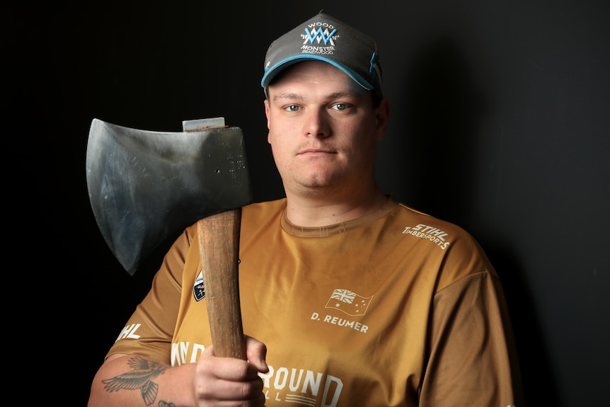 David holds an axe while wearing a blue and grey hat and brown t-shirt.
