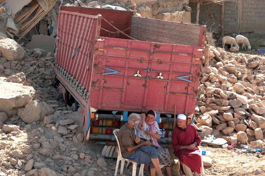 Three people sit on chairs in front of truck surrounded by debris 