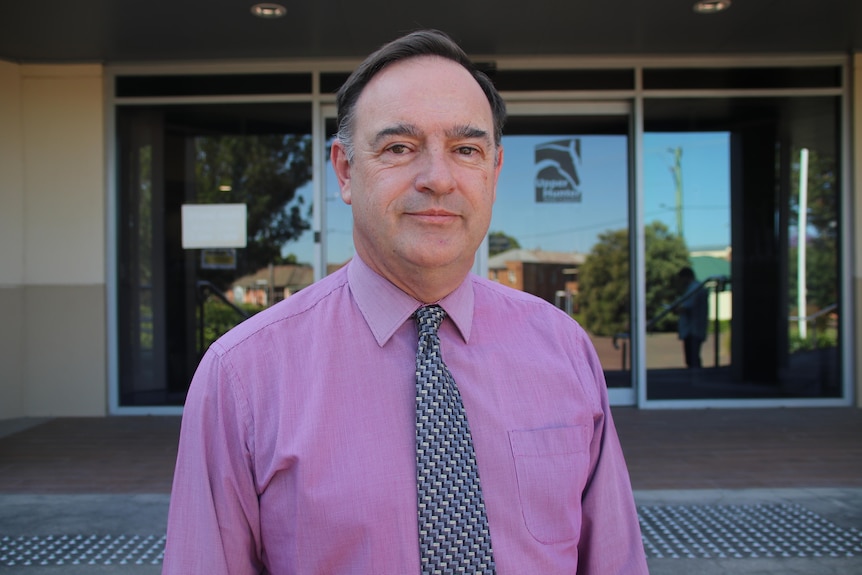 A middle-aged man with short, dark hair, wearing a suit and tie and standing in front of an office building.