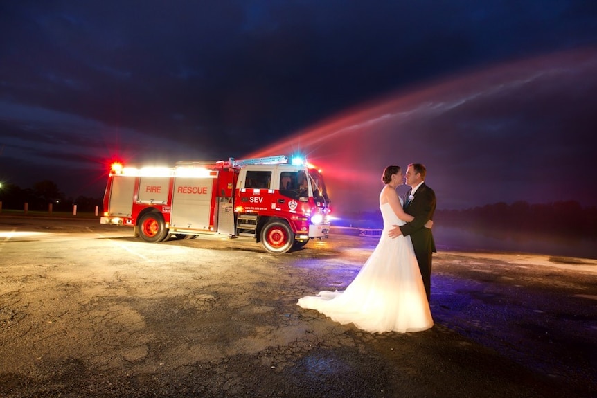 A bride and groom stand in front of a red fire truck with its lights on at night.