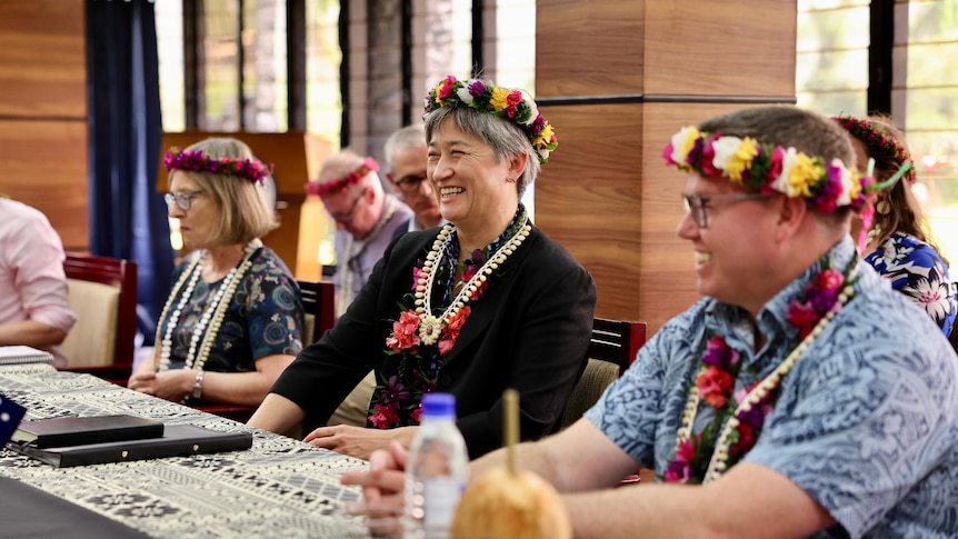 Penny wong in a floral head wreath and necklace sits at a table surrounded by others in similar garb. 