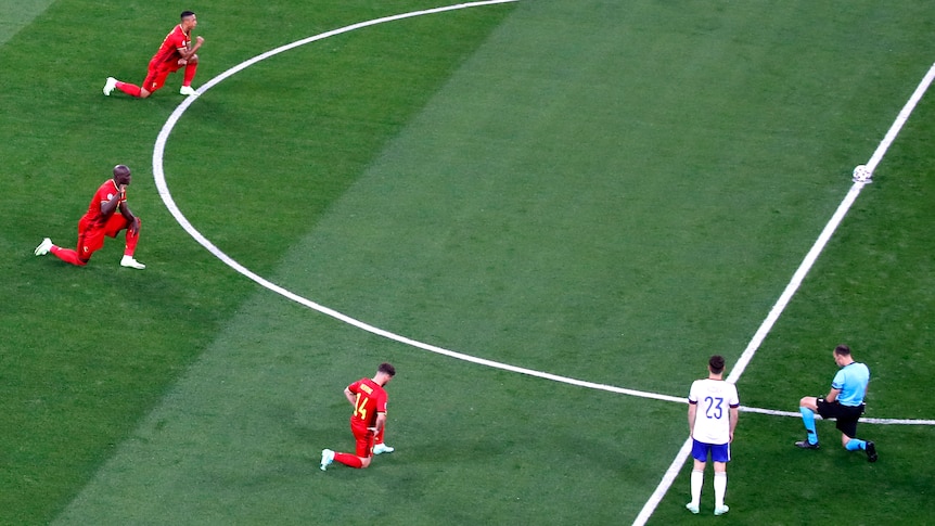 Four Belgium players, in red, and one referee, in blue, kneel as a player in white stands 