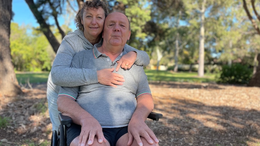 Graham in his wheelchair near a suburban park, with his wife Helen wrapping her arms around him.