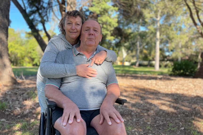 Graham in his wheelchair near a suburban park, with his wife Helen wrapping her arms around him.