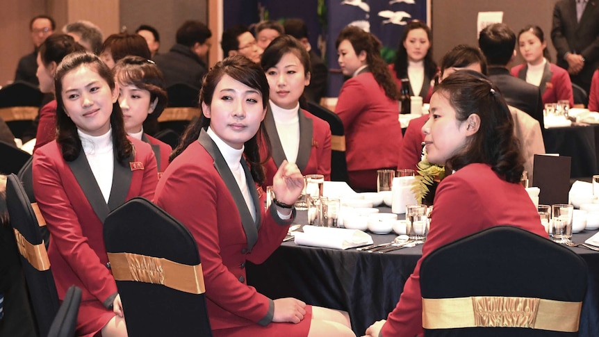 North Korean women wearing black-trimmed red blazers sit at banquet tables.
