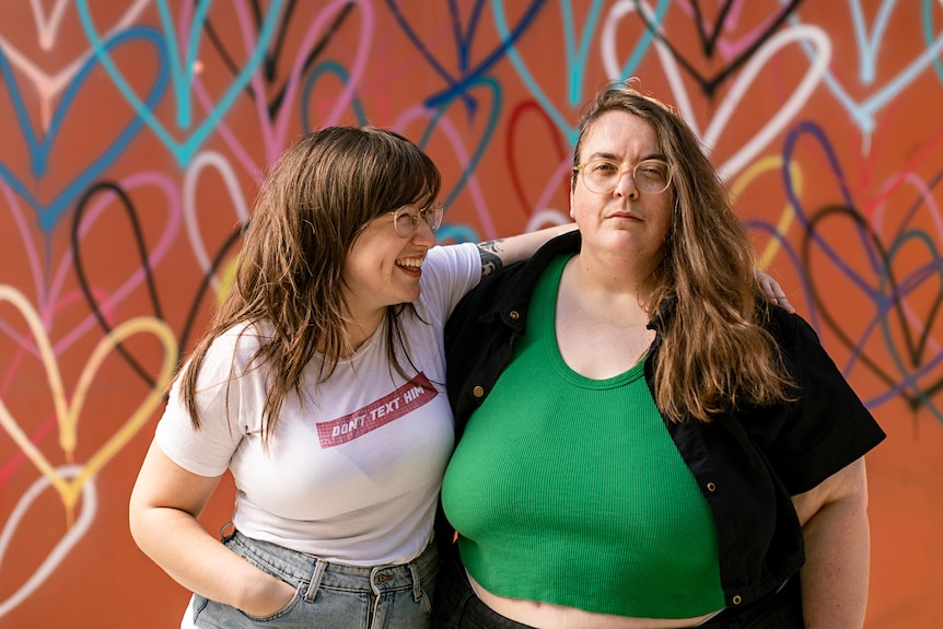 Freya, left, looks at Bec as she stands with her arm draped over her. Bec looks to the camera with a faint smile.