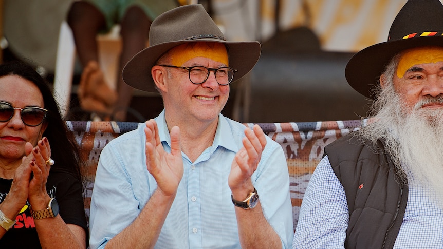 Anthony Albanese, wearing a brown akubra hat and blue shirt, sits with a woman and man clapping