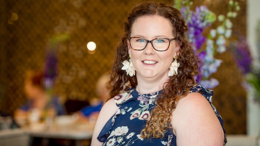 A profile image of a woman with long, curly hair and glasses, standing in a restaurant.
