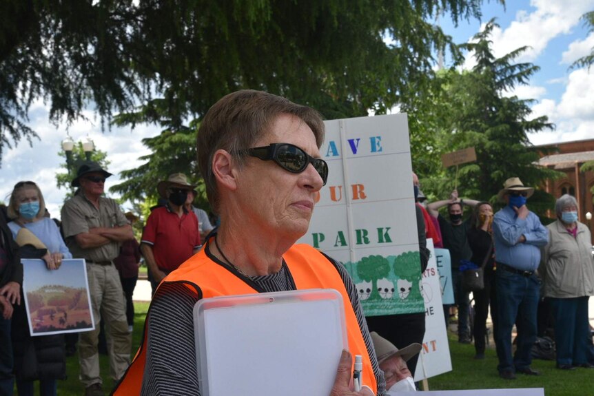 A woman wearing sunglasses and hi vis stands in front of protestors with a sign that reads "save our park".