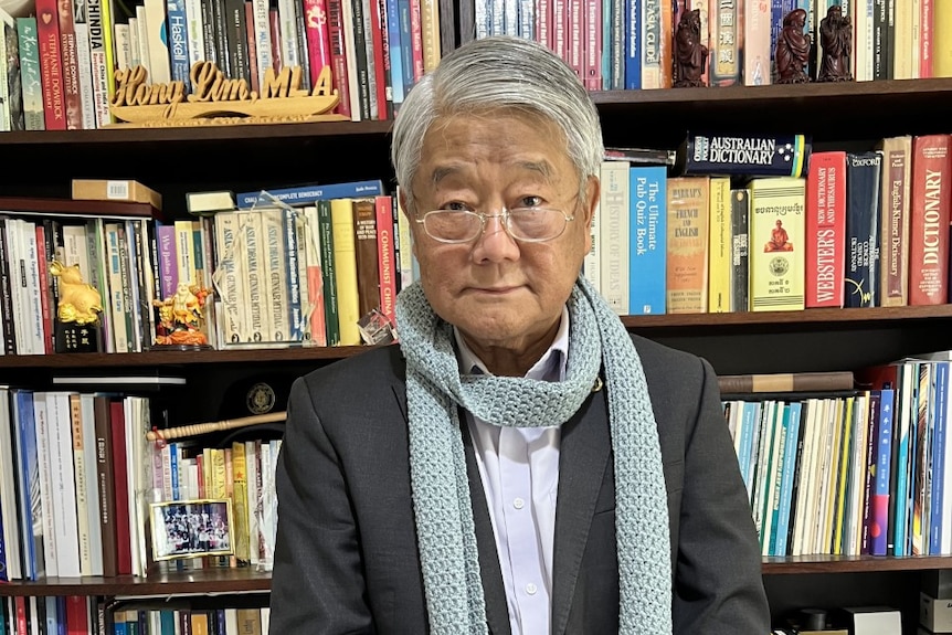 A man while white grey hair and glasses wearing crochet blue scarf in front of bookshelf