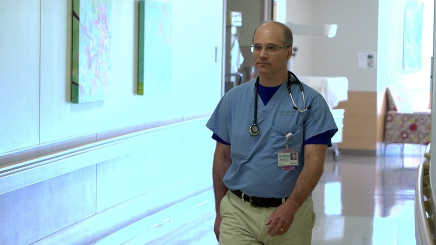 Dr Aaron Chidekel walks to the left of the image. He wears hospital scrubs on his upper half and has a stethoscope around neck.