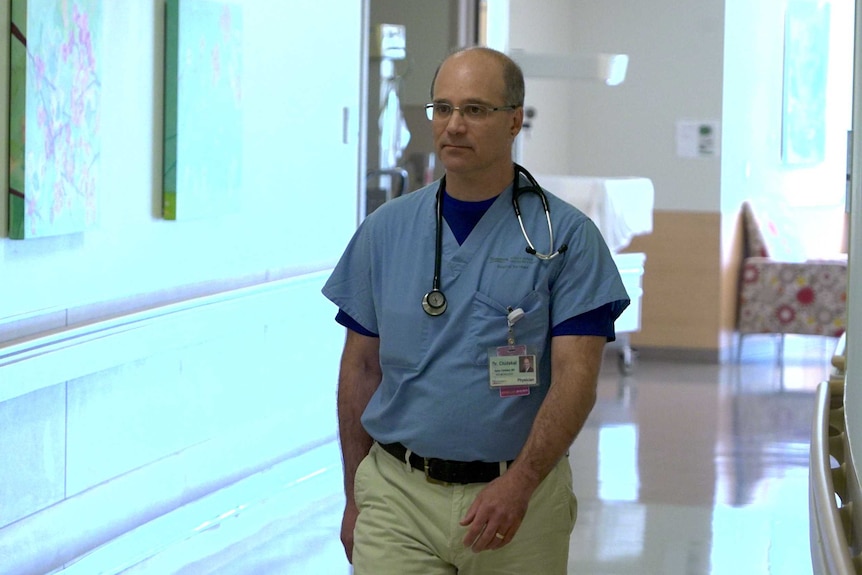 Dr Aaron Chidekel walks to the left of the image. He wears hospital scrubs on his upper half and has a stethoscope around neck.