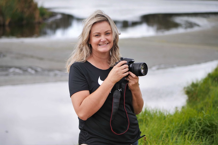 A smiling blonde woman in a black t-shirt holds a camera in front of a grass-edged lake