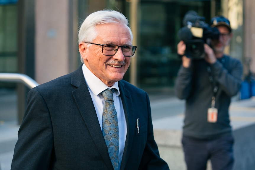 Duncan Ord wears a black suit and white shirt with a tie and glasses, outside a city building, with a TV camera crew in the back