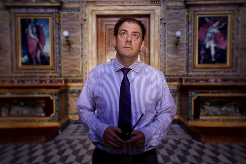 Ben Harnwell stands in a room with a tiled floor and ornate walls with paintings, looking up, as light shines on his face