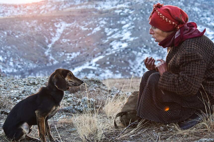 A woman in checked jacket with head wrapped in red scarf sits and with small black dog in remote and rocky terrain with snow.