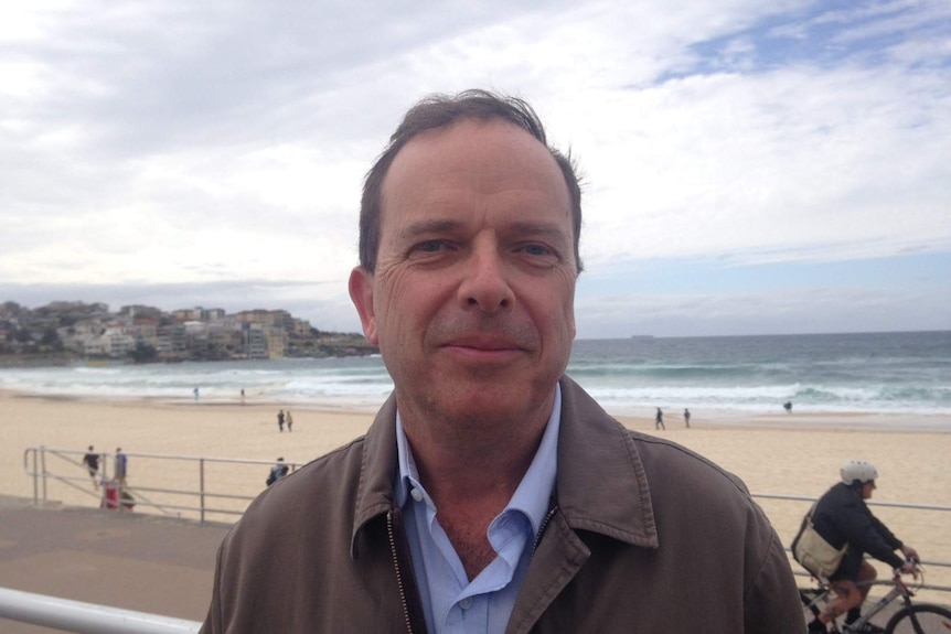 A man in a jacket and dress shirt standing in front of a beach.