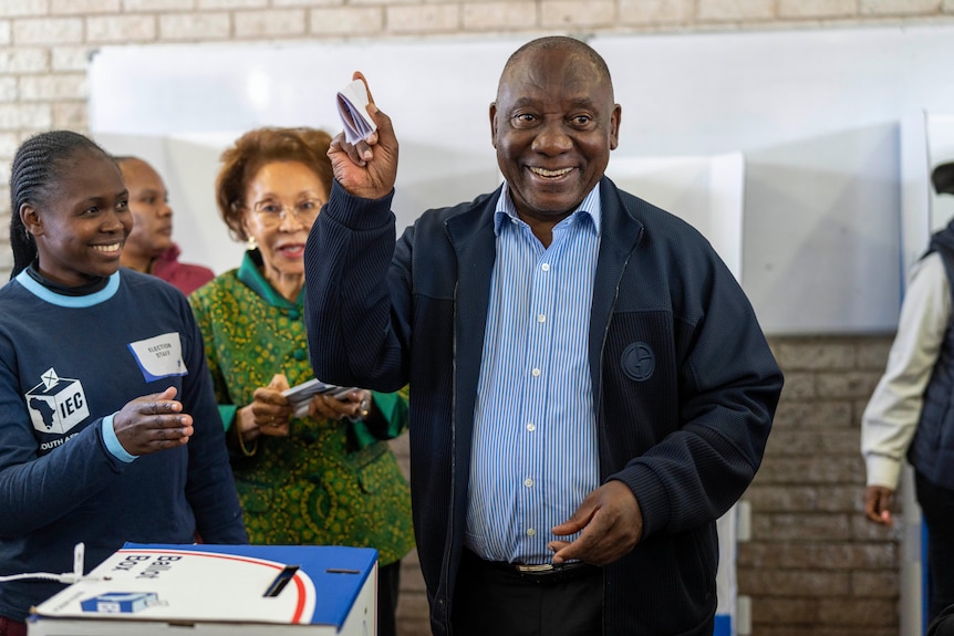 An older South African man in a casual jacket smiles as he goes to place a folded piece of paper in a ballot box.