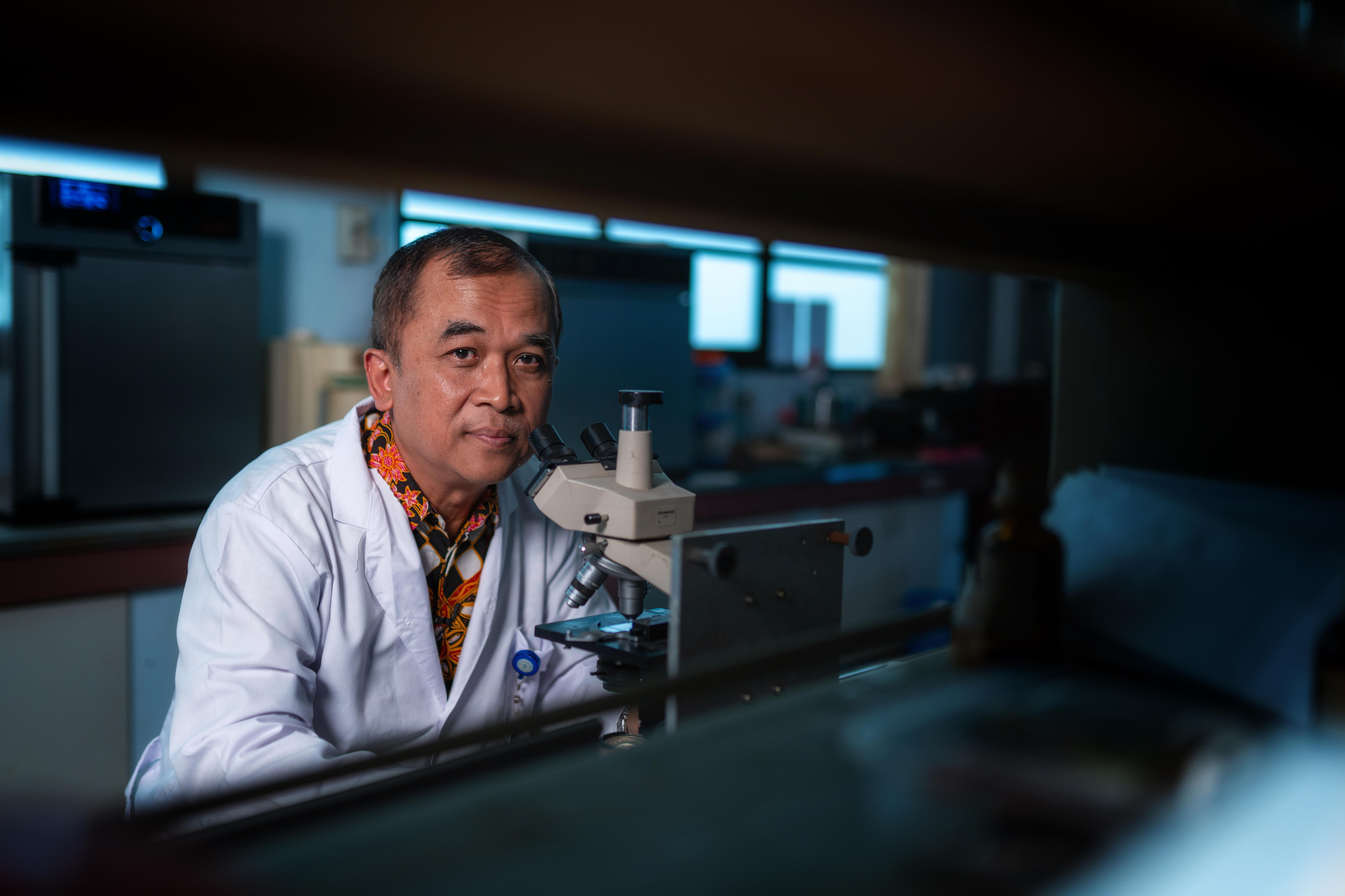 A man wearing a white lab coat over his orange and black patterened shirt sits in front of a microscope