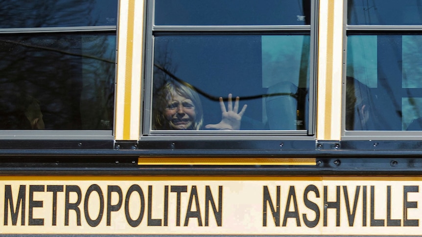 A little girl cries and places her hand on the window of a yellow school bus.