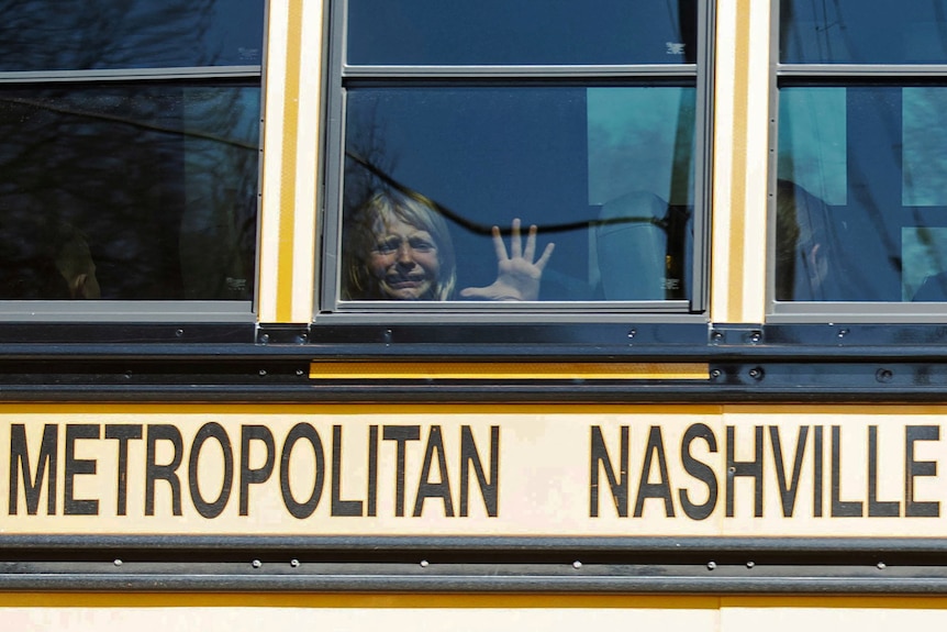 A little girl cries and places her hand on the window of a yellow school bus.
