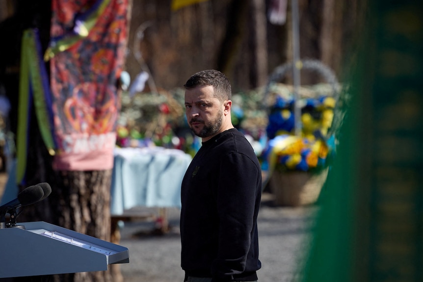 Zelenskyy looks down the camera lens in front of floral tributes