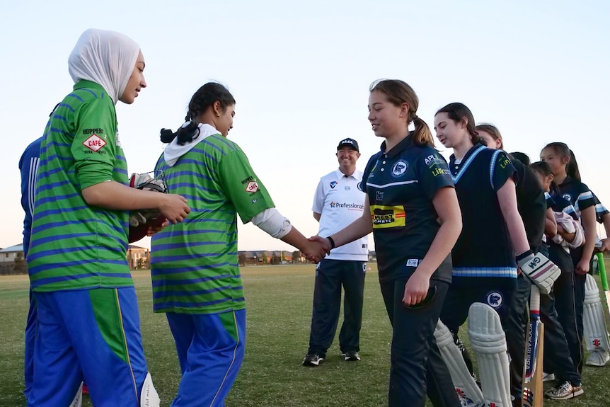 Two rows of girls dressed in cricket tracksuits shake hands as they walk past one another.