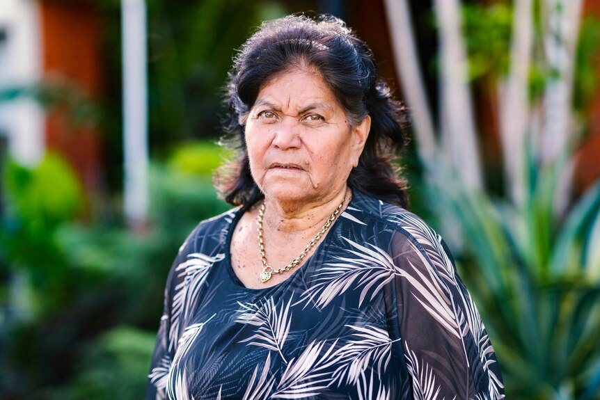 An older woman wearing a black and white top, standing in a garden. She has a sad look on her face.