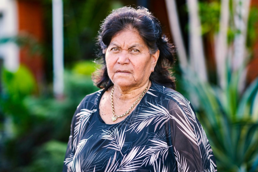 An older woman wearing a black and white top, standing in a garden. She has a sad look on her face.