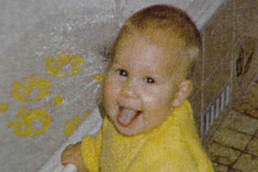 A baby girl wearing a yellow jumper holds on to the side of a bathtub
