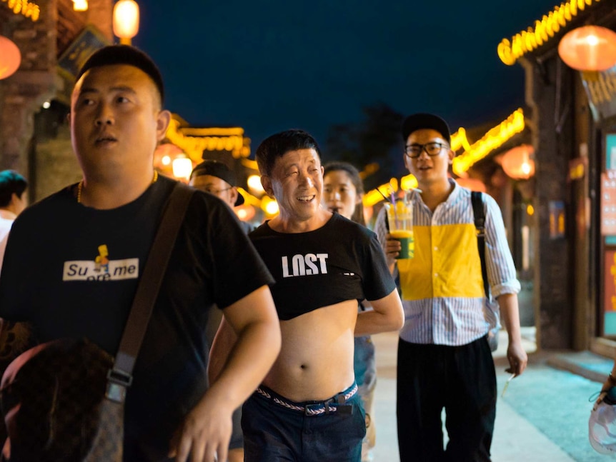 On a street at dusk, men walk down a marketplace with one at the centre exposing his midriff.