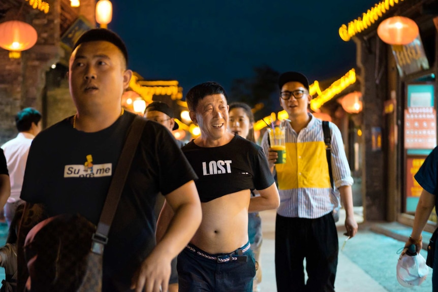 On a street at dusk, men walk down a marketplace with one at the centre exposing his midriff.