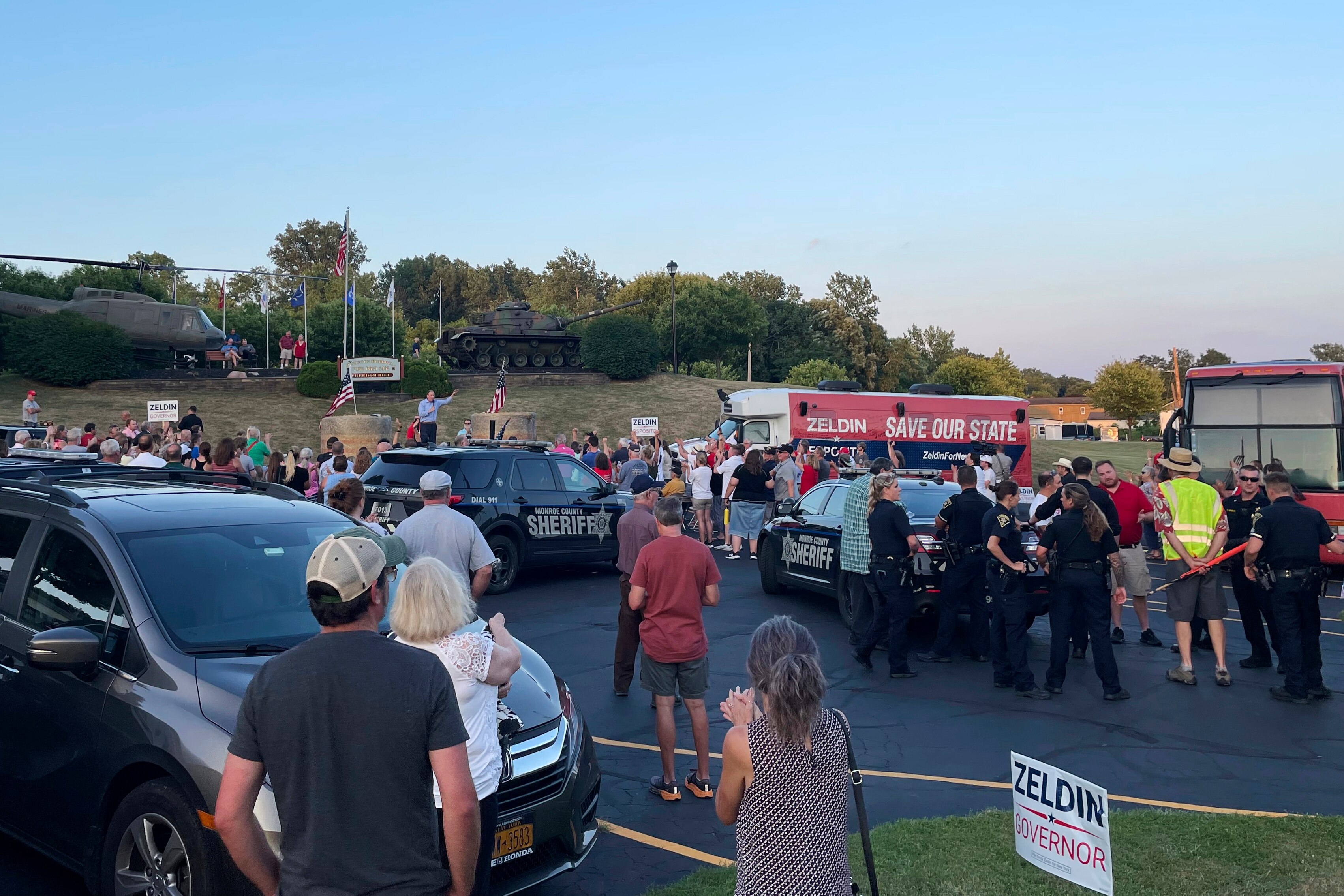 A man speaks to a crowd from a temporary podium to people and sheriffs scattered around a car park