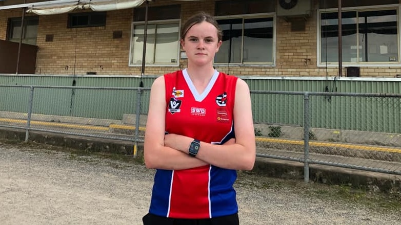 A teenage girl in a red guernsey drinks water at a team huddle.