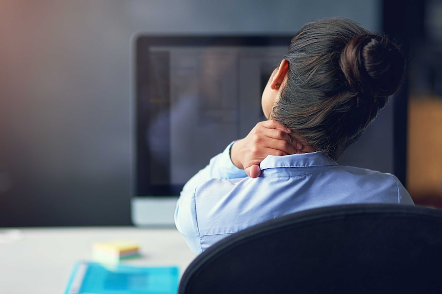 A woman sits at a computer, holding her neck.