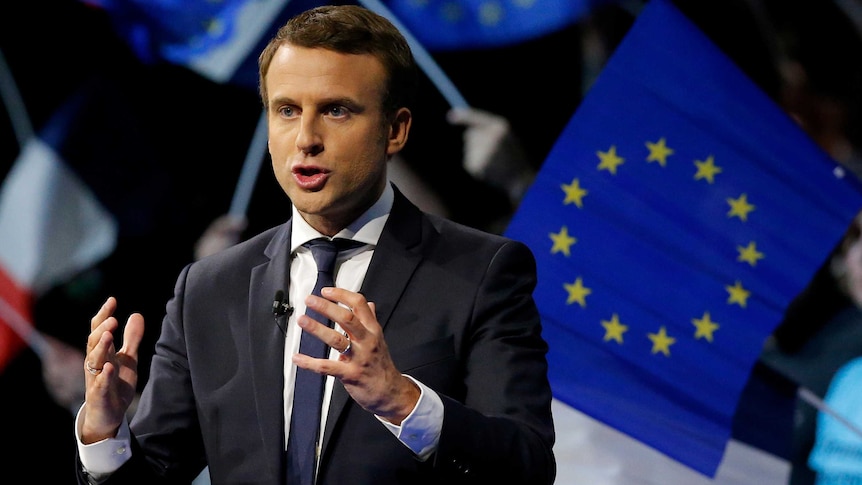 Emmanuel Macron speaks to a crowd with the flag behind him