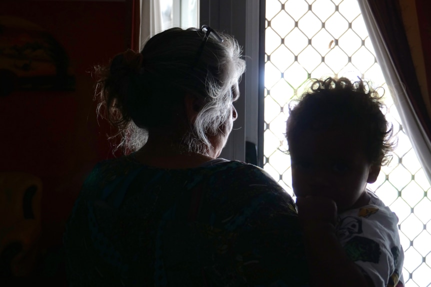 A woman with silver hair holding a baby looks out at a window. They are silhouettes.