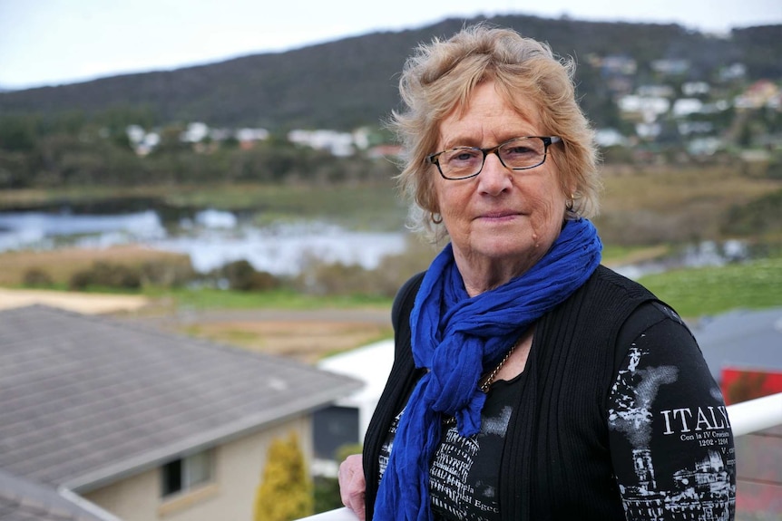 A woman with glasses, blonde hair, a blue scarf looks at the camera. In the background is a lake and a hill.