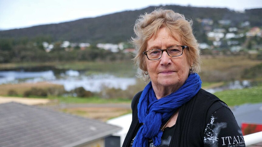 A woman with glasses, blonde hair, a blue scarf looks at the camera. In the background is a lake and a hill.