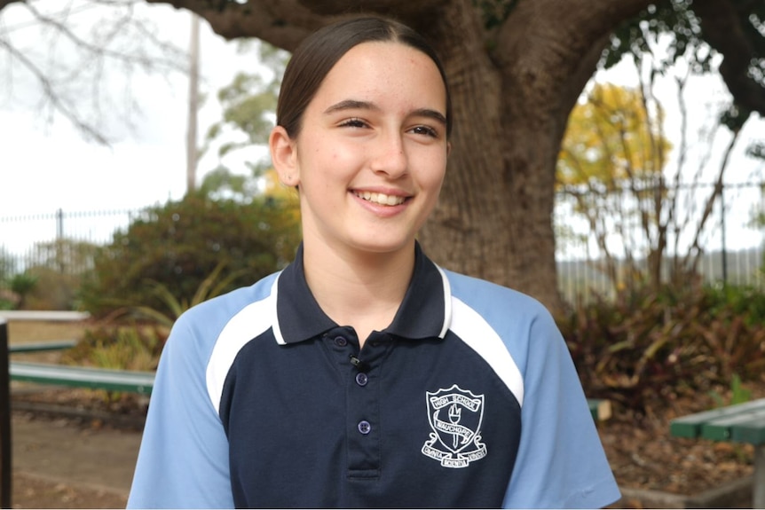 Young girl smiling wearing her school sport shirt in front of a tree