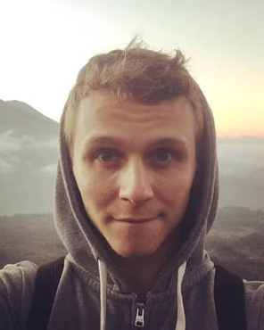 A young white man with a gray hood looks at the camera in front of a mountain landscape.