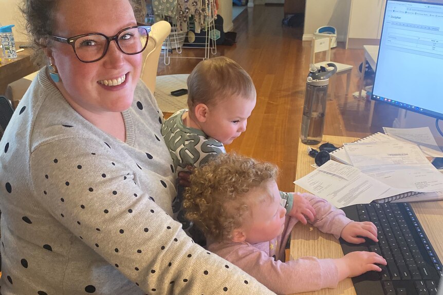 A woman sits at her desk at home in front of a computer, with two children in her lap.