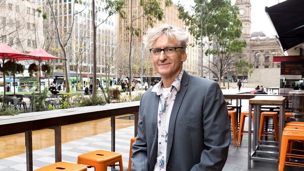 A man with wavy grey hair, wearing a jacket and glasses, sitting at an open-air cafe in Melbourne.