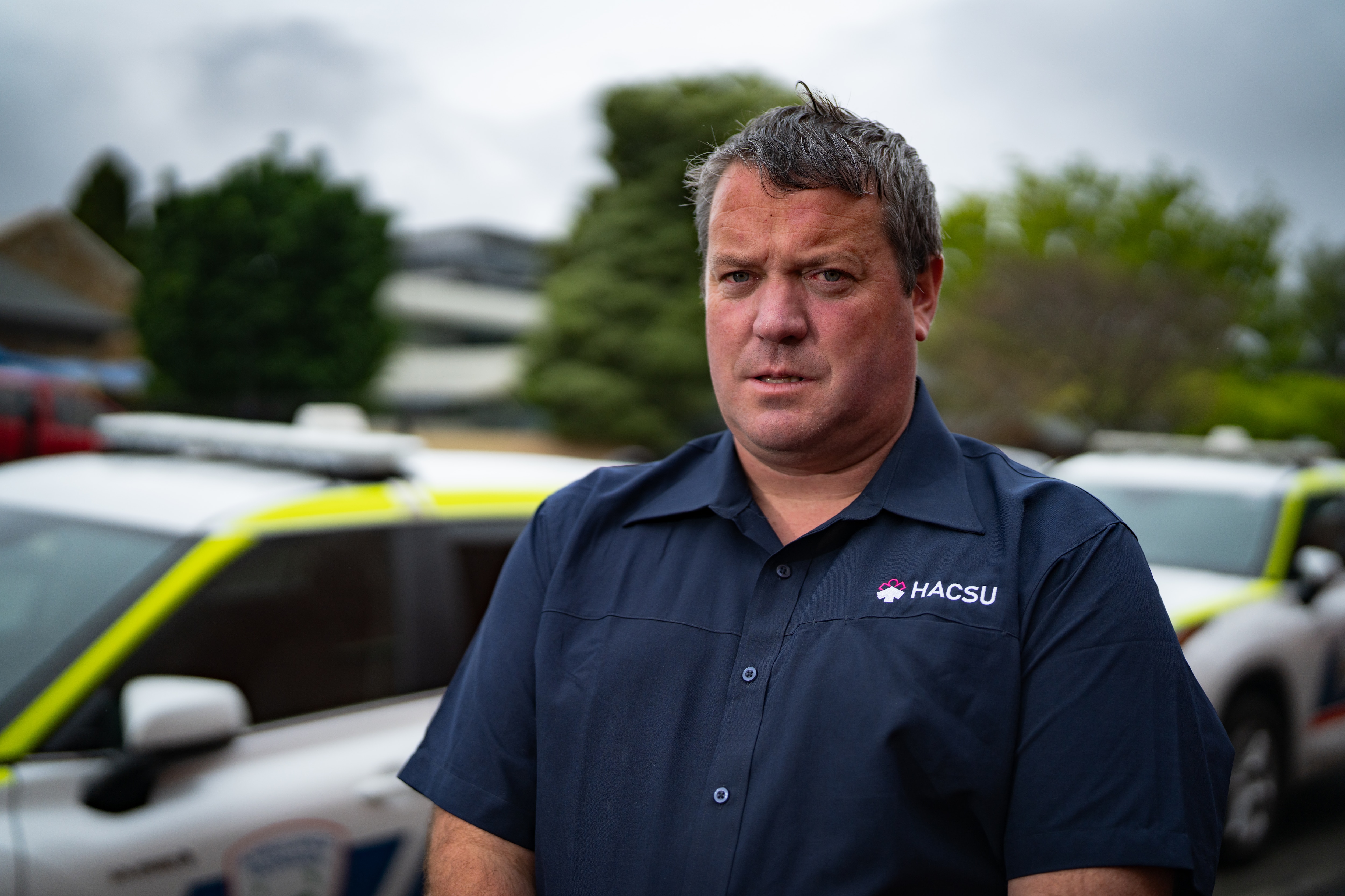 A man in a blue HACSU polo stands in front of an ambulance outdoors on an overcast day