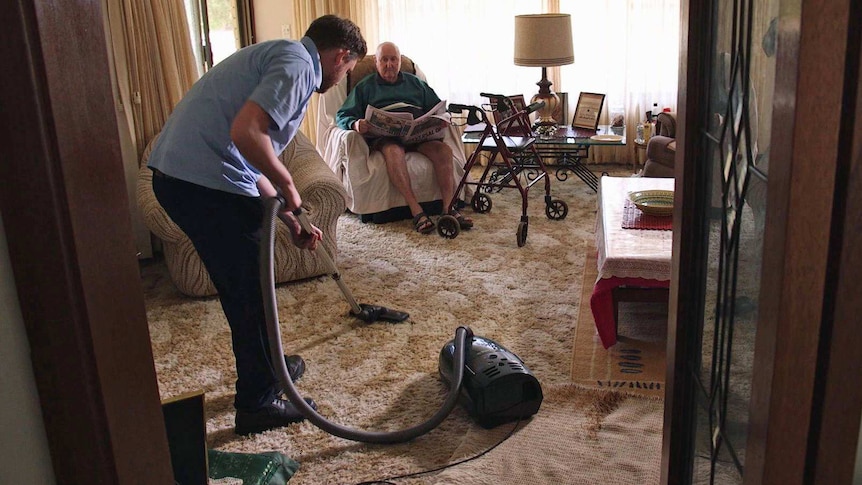 A man vacuums while an elderly man sits and watches