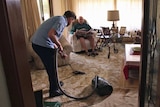 A man vacuums while an elderly man sits and watches
