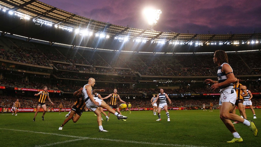 An AFL player kicks for goal in front of a crowd of players as flood lights in the background shine down on the grandstands