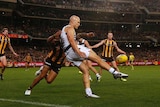 An AFL player kicks for goal in front of a crowd of players as flood lights in the background shine down on the grandstands