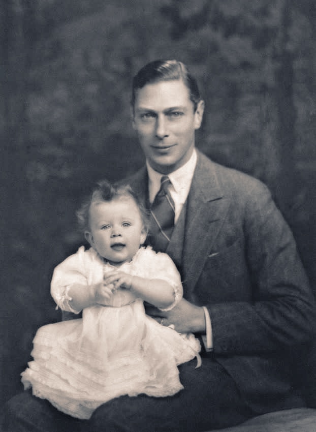 King George VI holds a baby Queen Elizabeth on his knee in a black and white photograph.